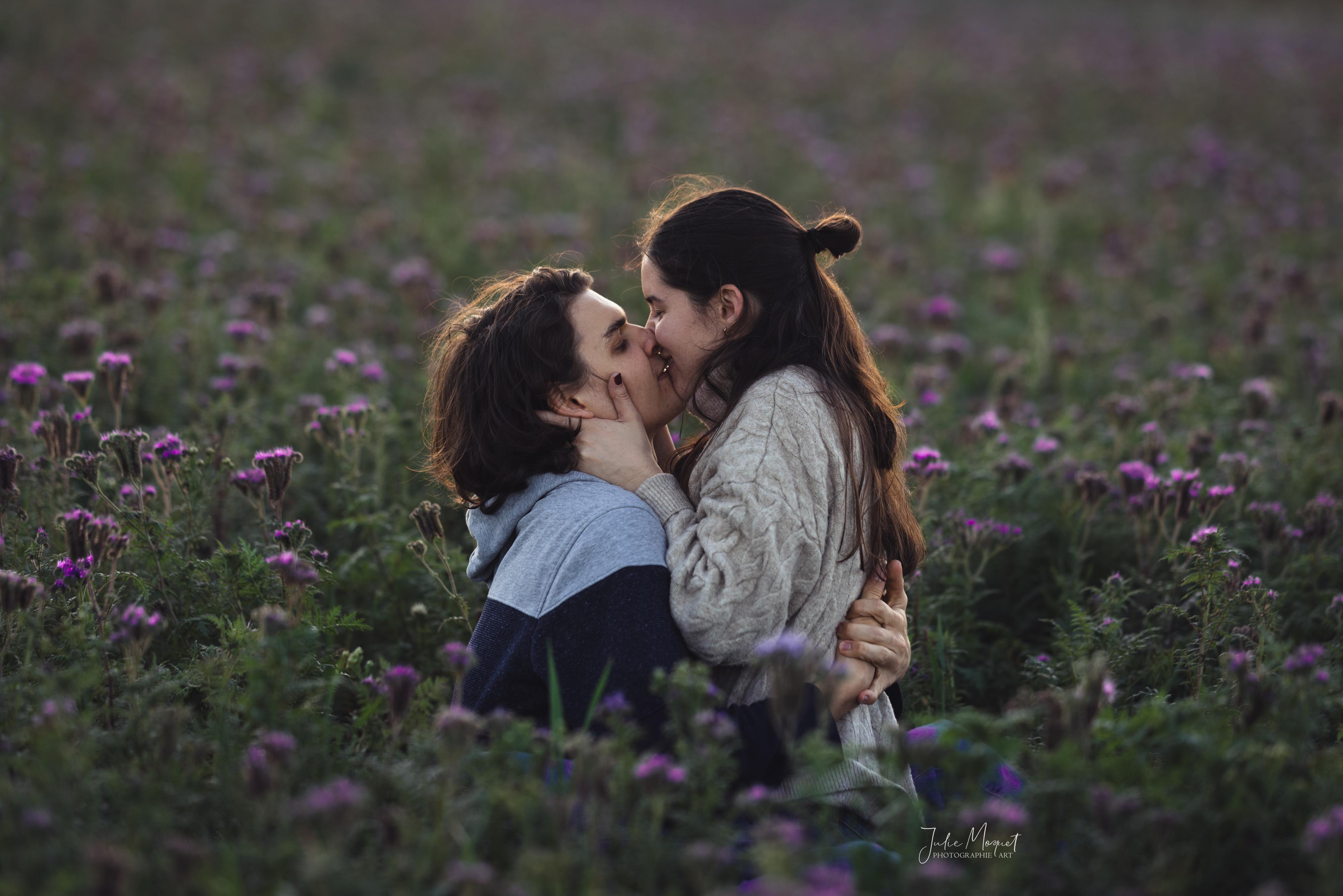 Ein junges Paar (Mann und Frau) aufeinander sitzend und sich küssend inmitten eines lila Blumenfeldes.