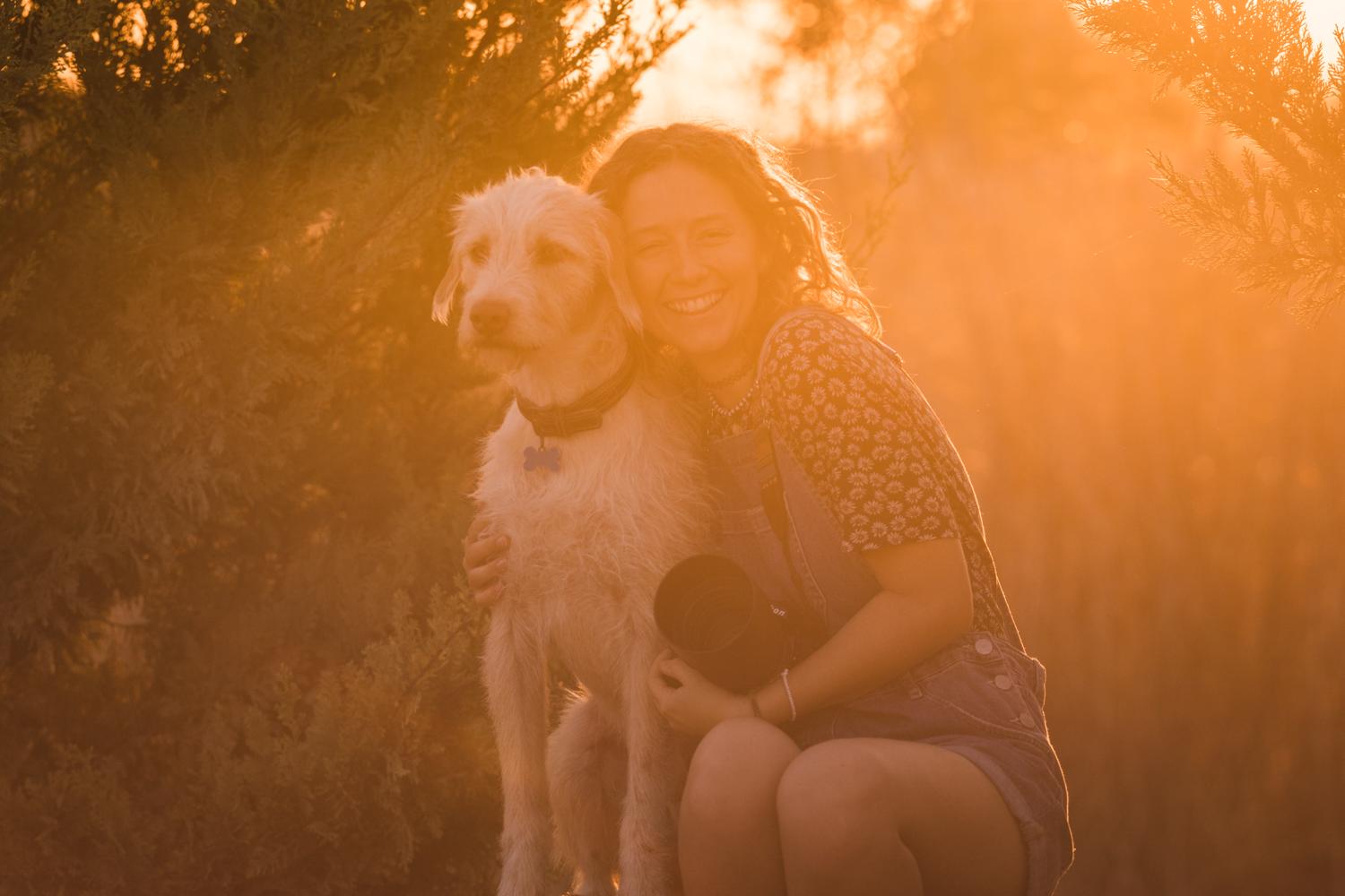 Die Fotografin Julie Moquet im Gegenlicht lehnt sich an einen  Hund, lächelt in die Kamera, hält ihren Fotoapparat im Schoß.