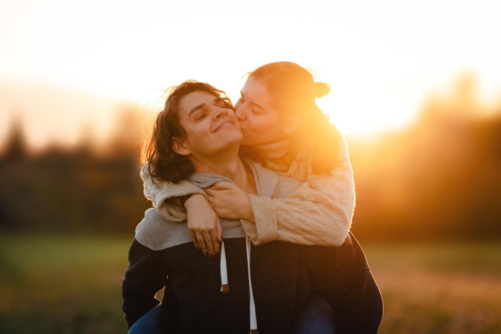 Portrait eines jungen Paares im Sonnenuntergang. Die Frau auf dem Rücken des Mannes gibt ihm einen Kuss auf die Wange.