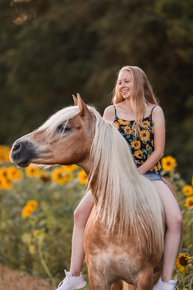 Portrait einer lächelnden jungen Frau auf ihrer Haflingerstute sitzend vor einem Sonnenblumenfeld im weichen Abendlicht.