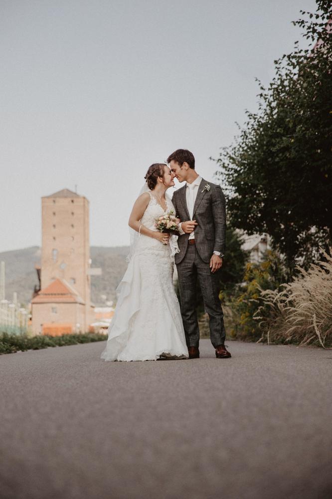 Rechts ein heterosexuelles Brautpaar Nase an Nase auf einem Weg. Links im Hintergrund ein Turm aus Backstein.