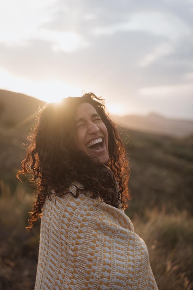 Eine dunkelhaarige Frau mit Locken im Sonnenaufgang nach hinten zu Kamera drehend, ausgiebig lachend in hügeliger Landschaft.