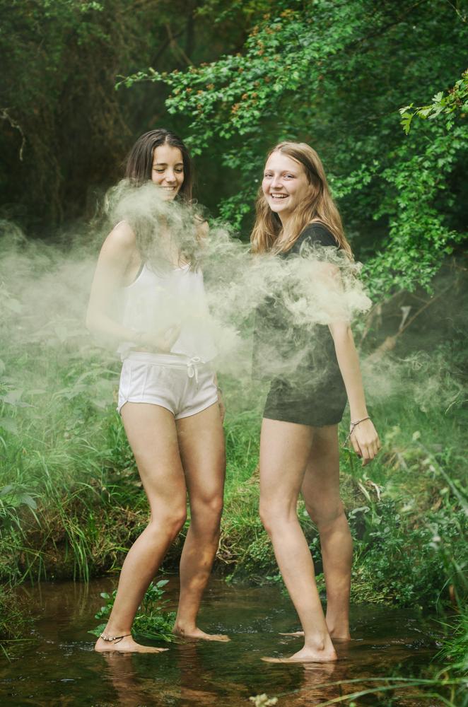 Zwei Mädchen in einem dicht bewachsenen Bach stehend mit einer Rauchbombe in der Hand. Um sie herum gelblicher Rauch/Nebel.