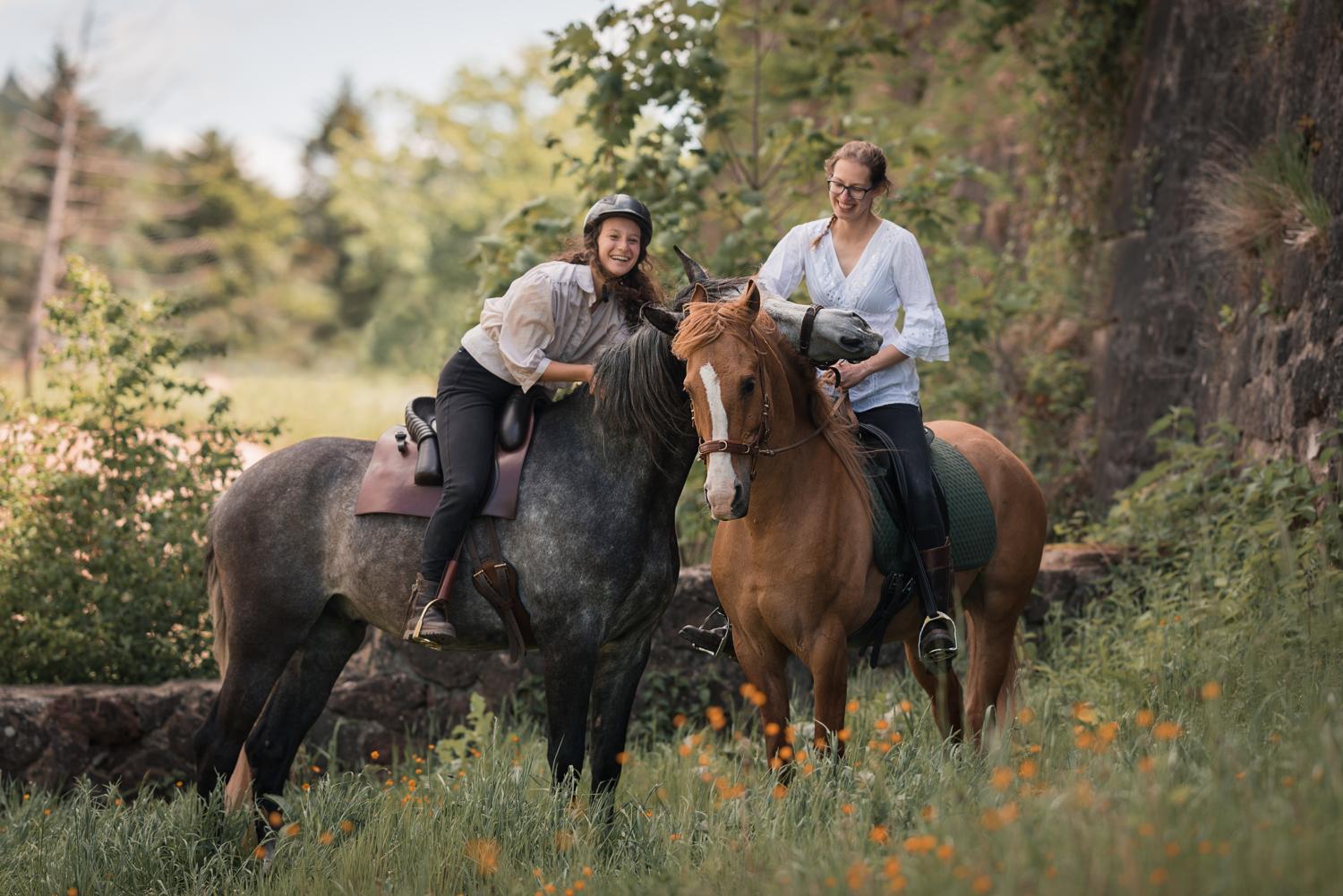 Die Reiterinnen Nativehorses auf zwei Pferden neben einer alten Burg. Das eine Pferd legt den Kopf über den Hals des anderen.