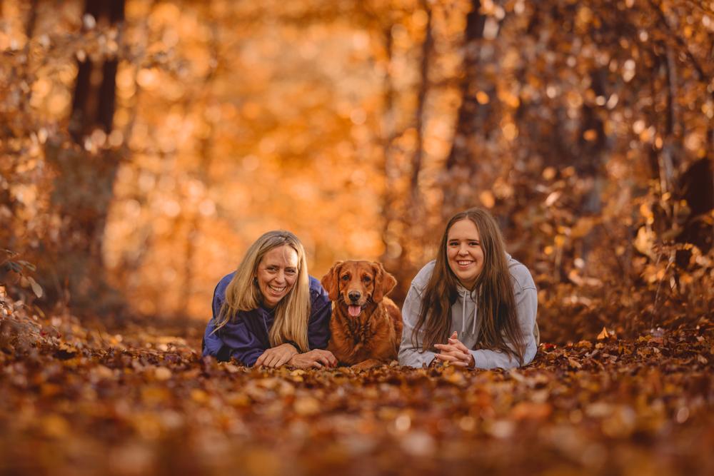 Zwei Frauen und ein Golden Retriever auf dem laubbedeckten Waldboden liegend und in die Kamera lachend im goldenen Herbst.