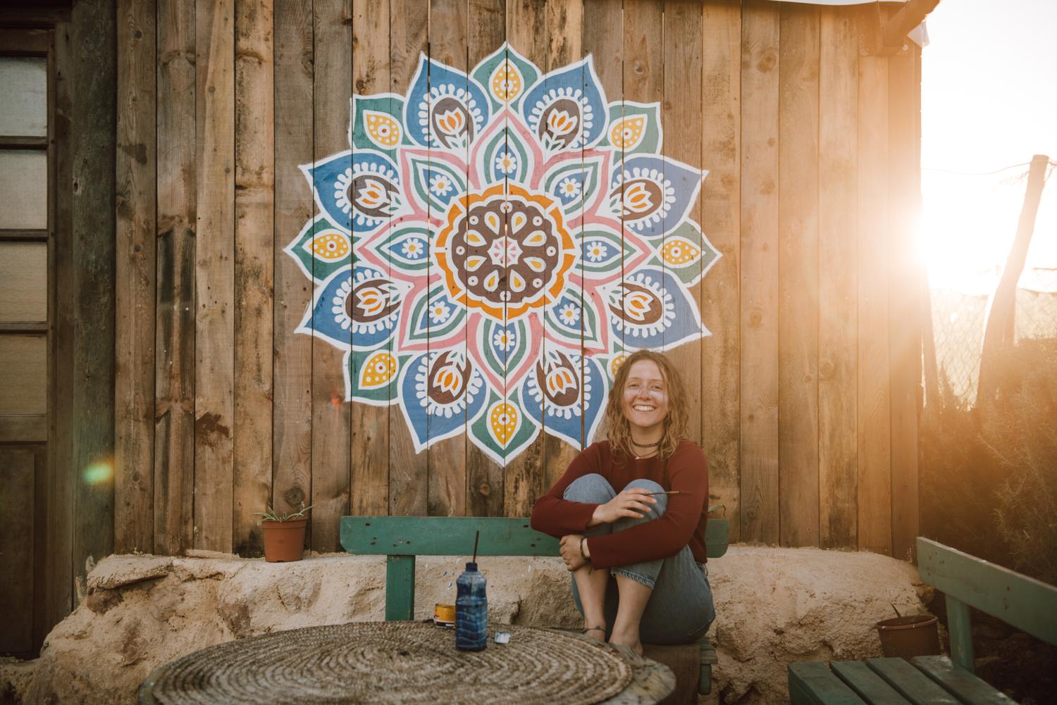 Buntes handgemaltes Mandala auf einer Holzwand darunter sitzt die Künstlerin. Hinter dem Gebäude geht die Sonne unter.